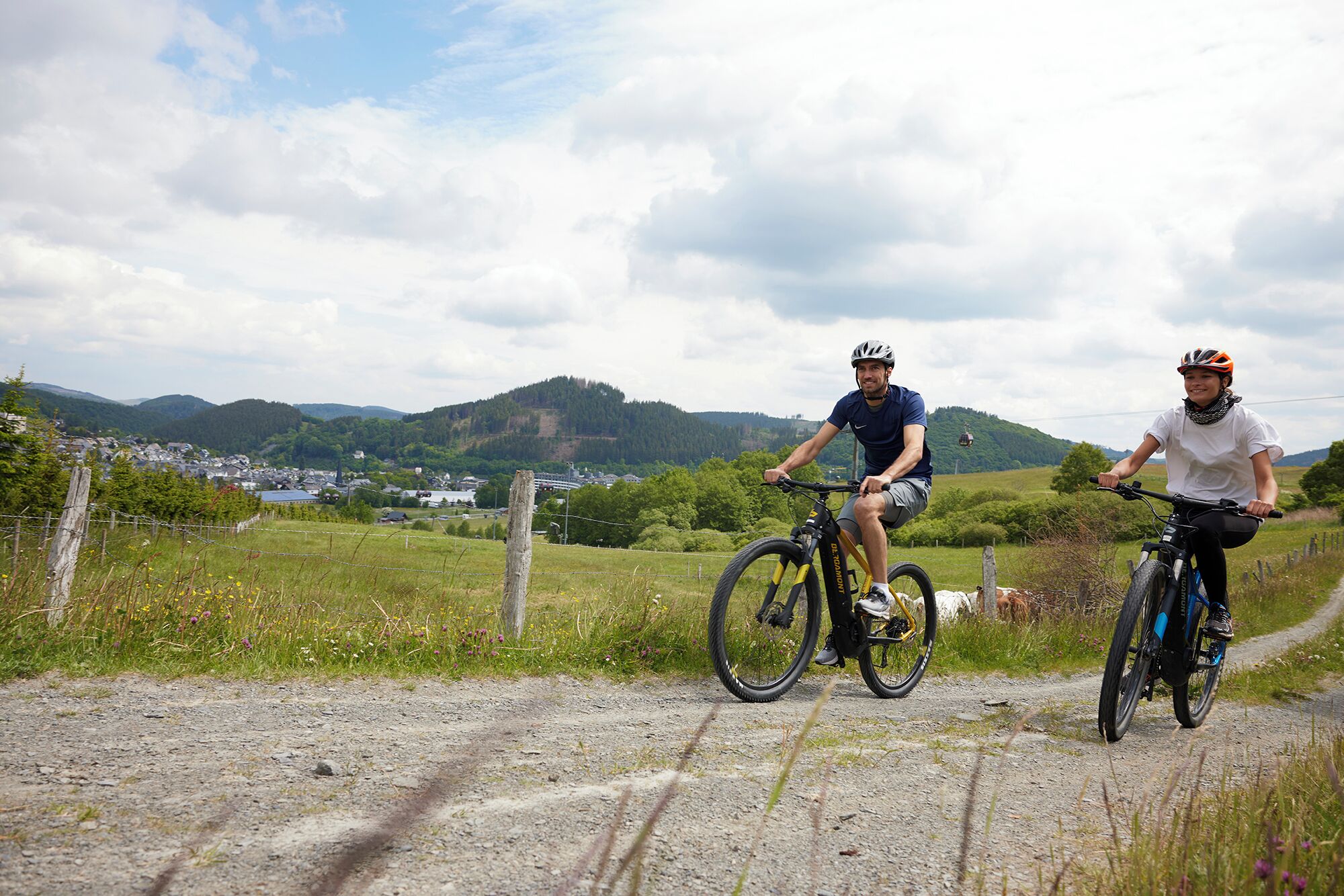 Two cyclists on a gravel path with a scenic view of a town and hills in the background.