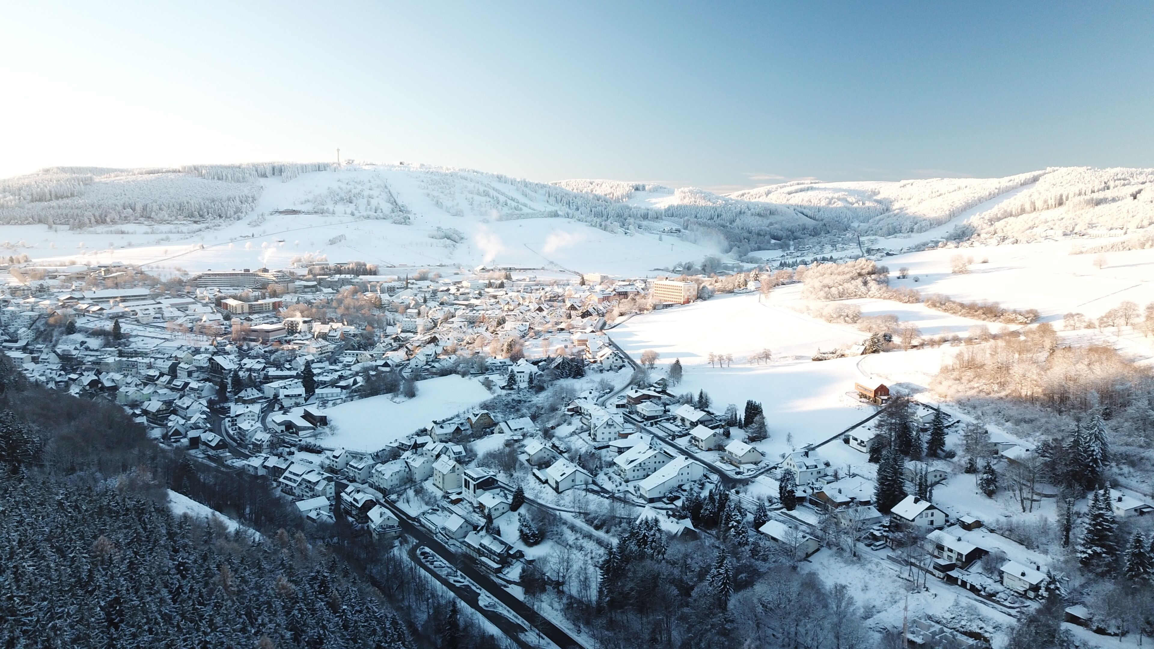 A snow-covered town with a mix of residential and commercial buildings, a central square, and surrounding hills.