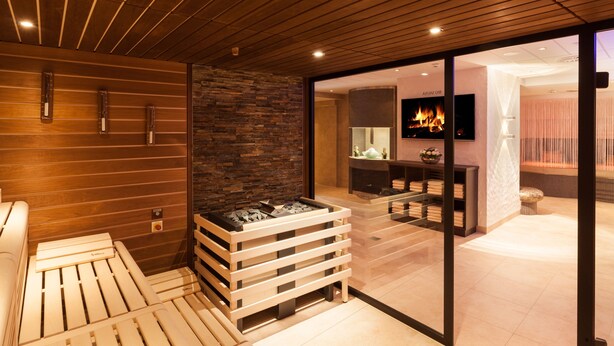 Ein moderner Wellnessbereich mit Holzbalkendecke, Steinwand und Kamin. Es gibt eine Sitzecke mit Fernseher und einem kleinen Tisch mit Handtüchern.