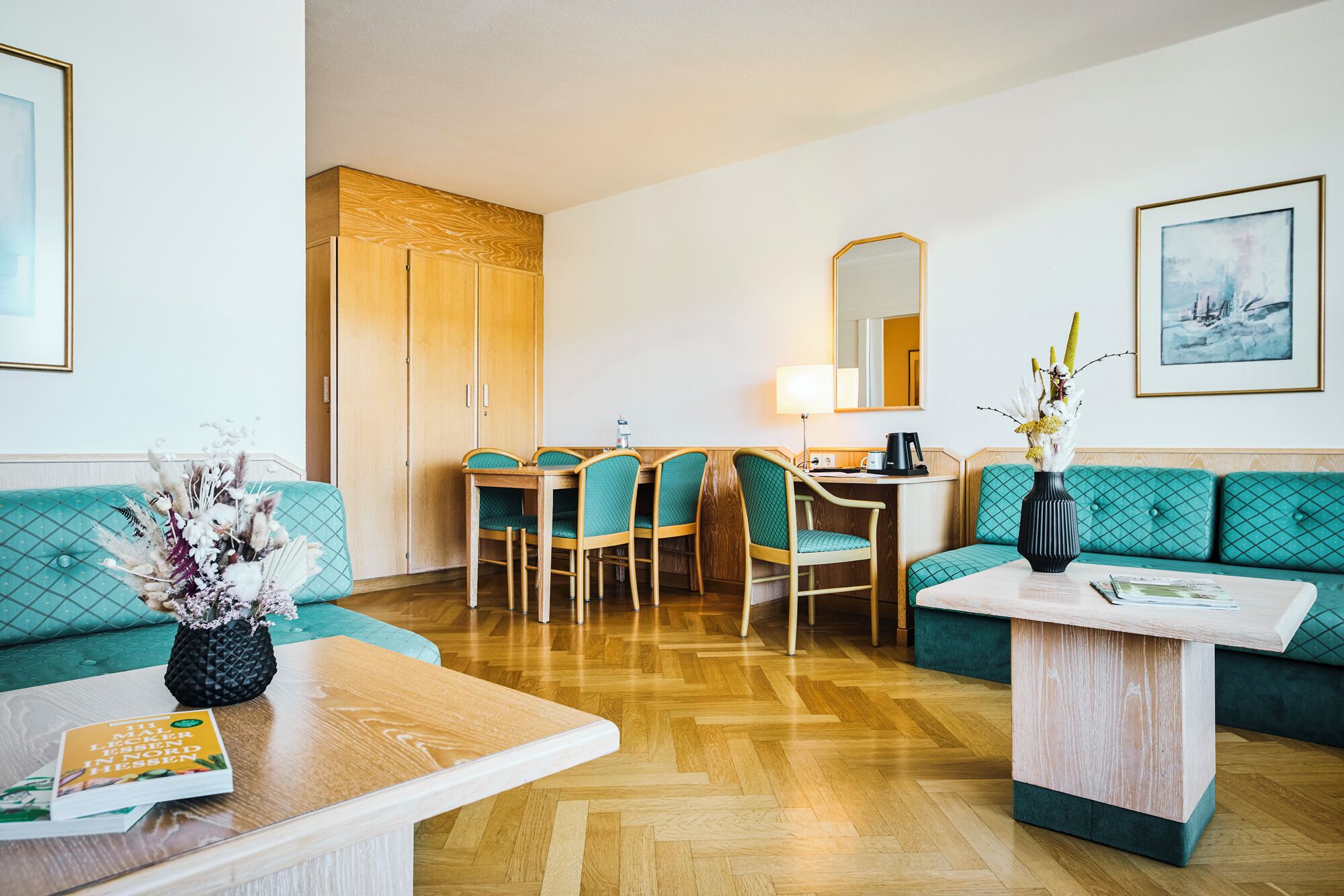A hotel room with a wooden floor, teal upholstered sofa, a wooden dining table with chairs, a small white table with a vase, and a mirror above the table.