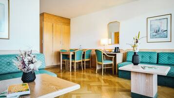 A hotel room with a wooden floor, teal upholstered sofa, a wooden dining table with chairs, a small white table with a vase, and a mirror above the table.