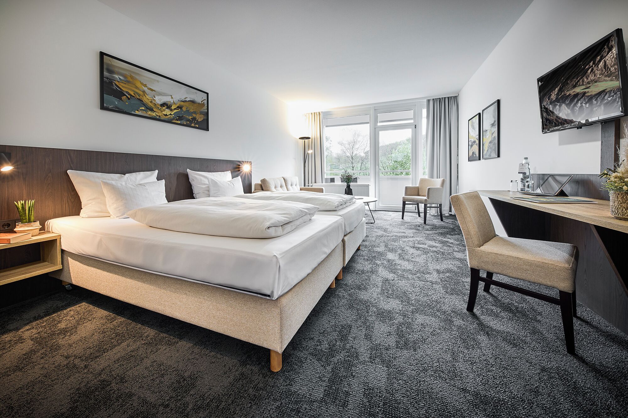 A hotel room with a large bed, a desk, and a chair. There is a window with curtains, a plant, and artwork on the walls.