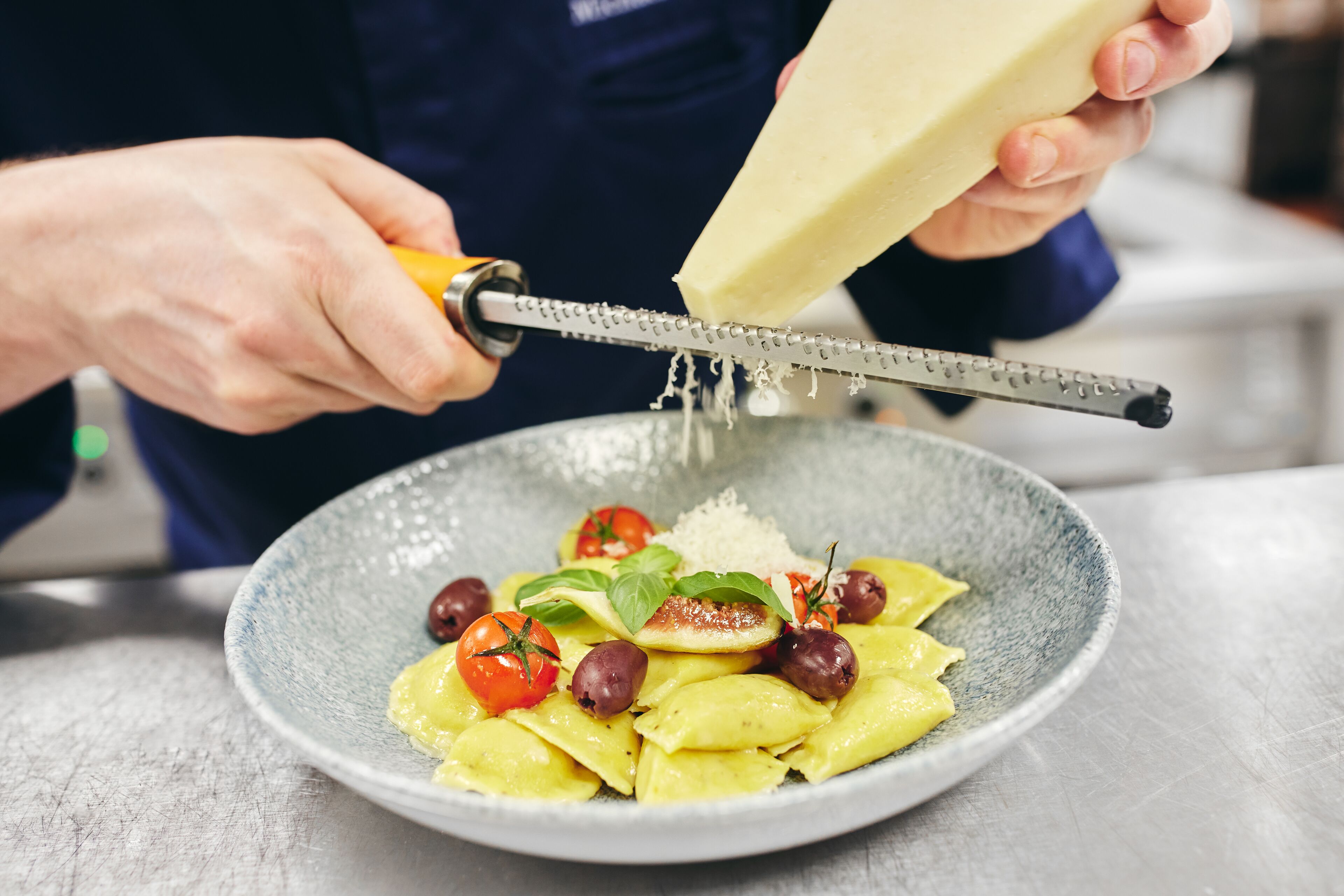 Eine Person reibt Käse über eine Pasta-Schale mit Tomaten und Oliven.