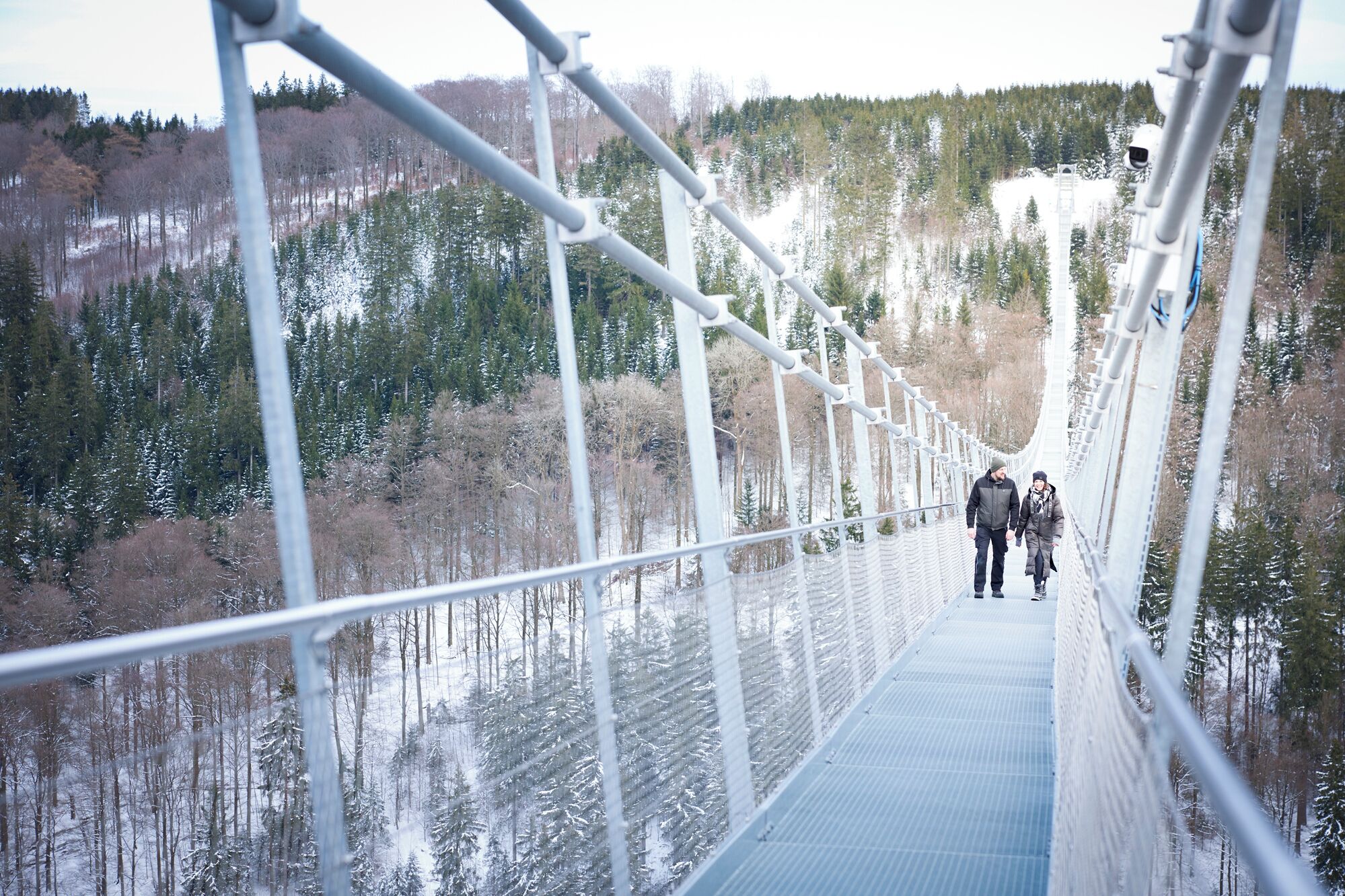 Un puente colgante con barandales de metal y un pasillo azul, rodeado de árboles nevados y un paisaje montañoso.