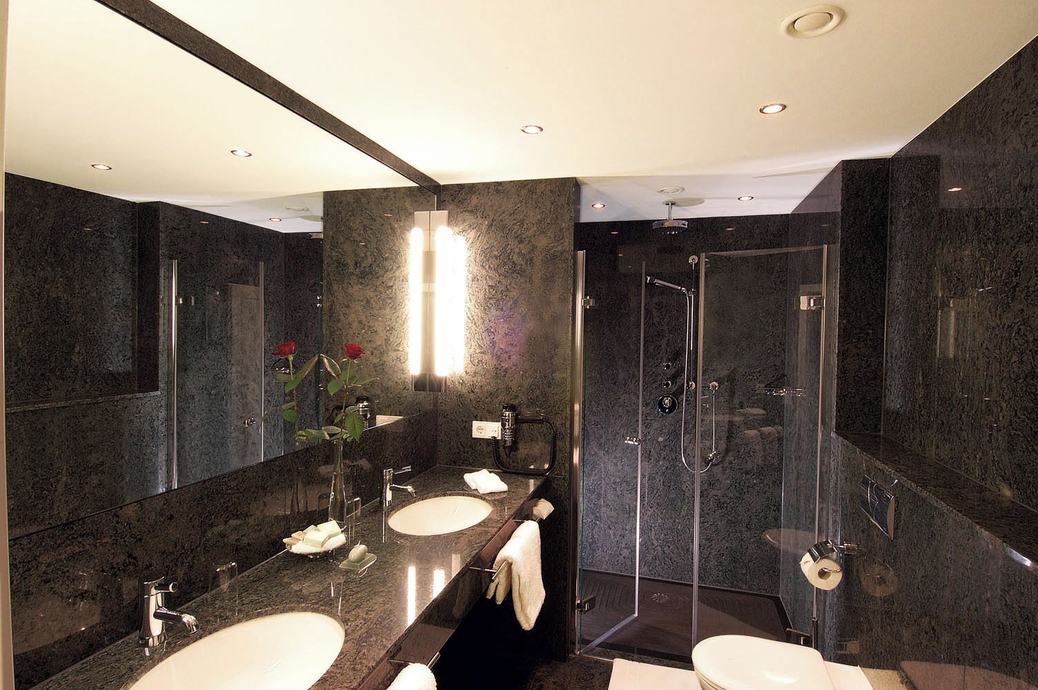 A modern bathroom with dark marble walls, a large mirror, two sinks, and a glass-enclosed shower.