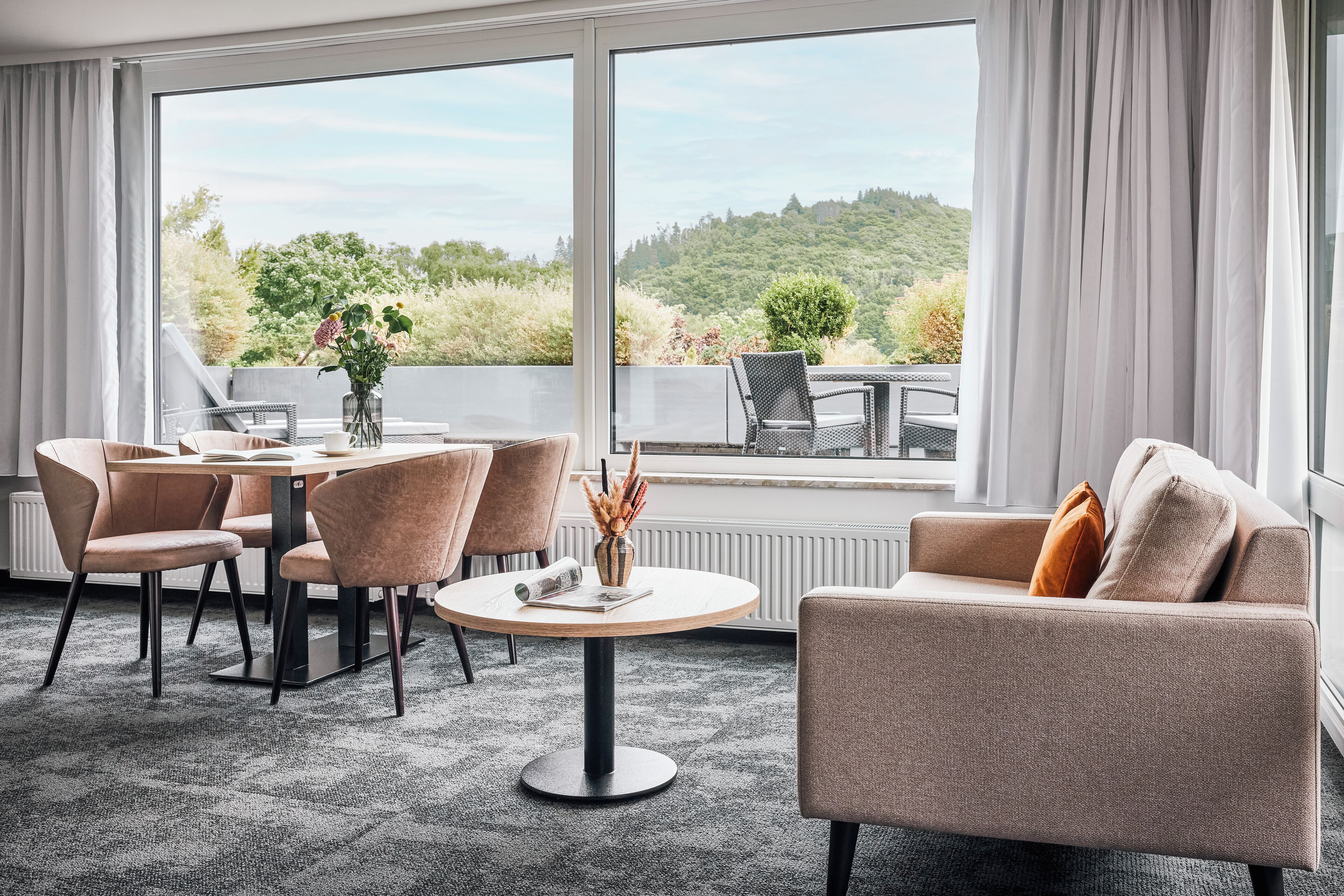 A modern hotel room with a round dining table, two sofas, and a large window offering a scenic view of greenery.