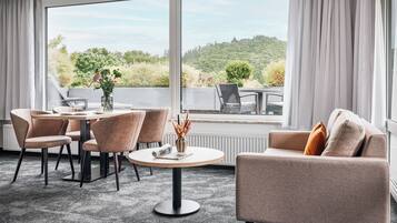 A modern hotel room with a round dining table, two sofas, and a large window offering a scenic view of greenery.