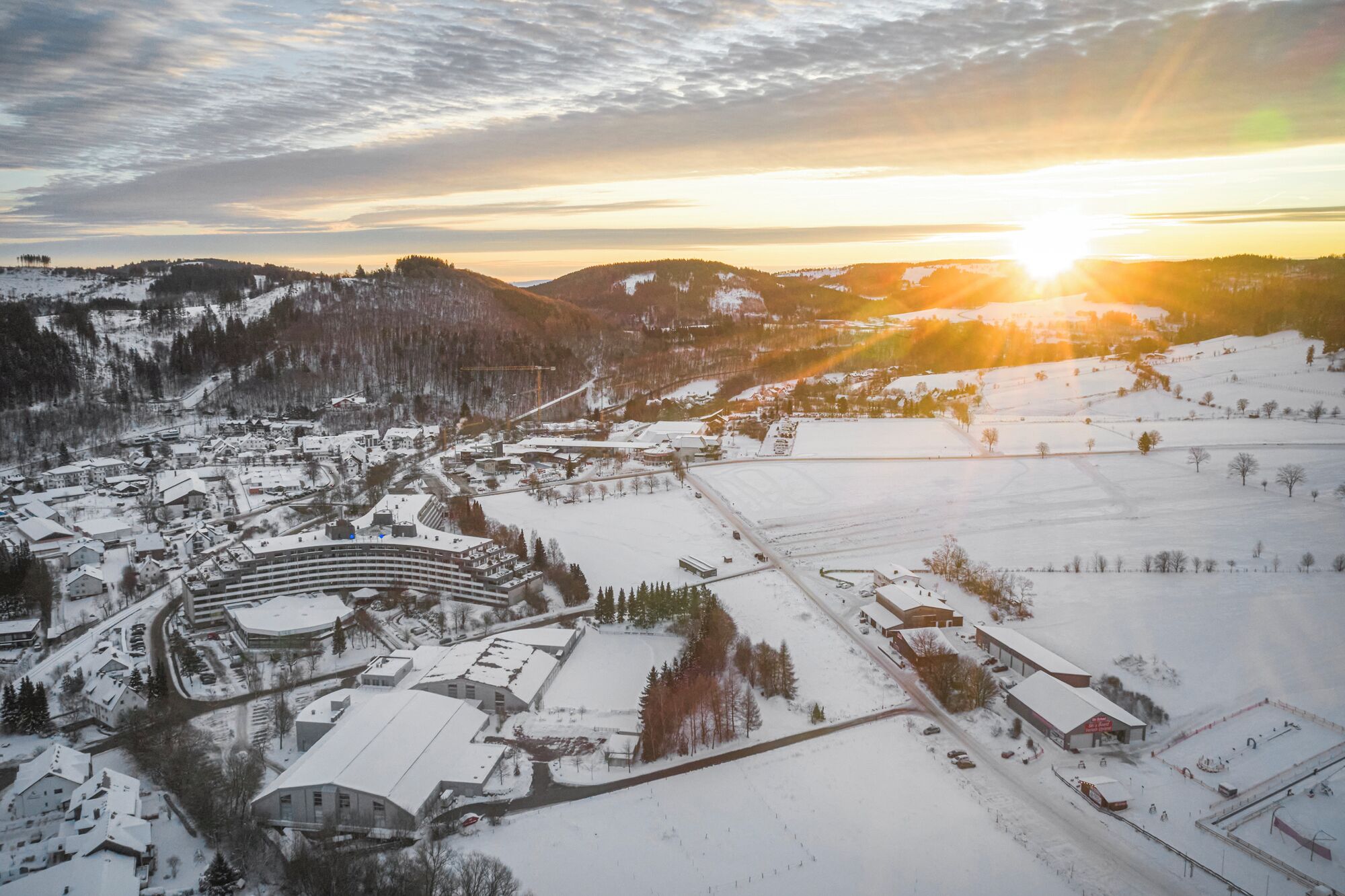 Une ville recouverte de neige, avec un imposant bâtiment circulaire, probablement un hôtel ou un centre de conférences, entouré d’immeubles résidentiels et commerciaux.