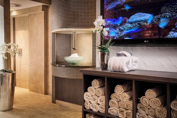 A modern bathroom with a large sink, a bowl of white flowers, a potted plant, and a TV displaying a nature scene. - Sauerland Stern Hotel (Willingen)