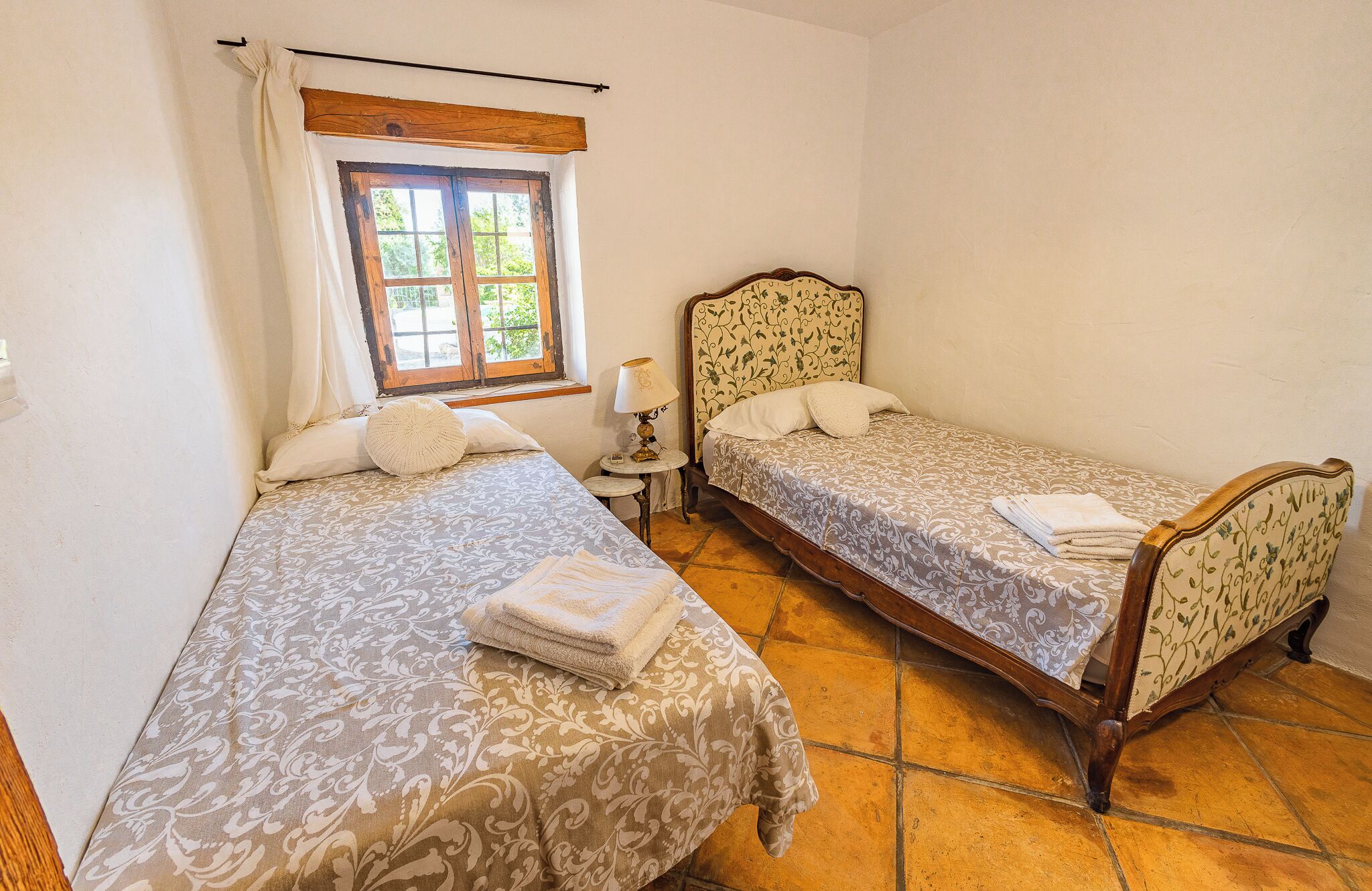 Quarto com duas camas de solteiro, janela com cortinas, mesa de cabeceira com abajur e piso de madeira.