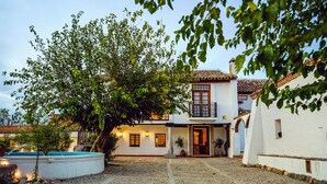 A courtyard with a swimming pool, a white building with a tiled roof, and a large tree.
