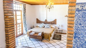 A bedroom with a wooden ceiling, a bed with a headboard, a chandelier, and a view through an archway.