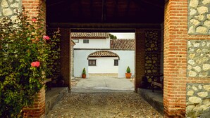 A courtyard with a cobblestone path leading to a white building with a tiled roof. There are potted plants and a bench in the courtyard.