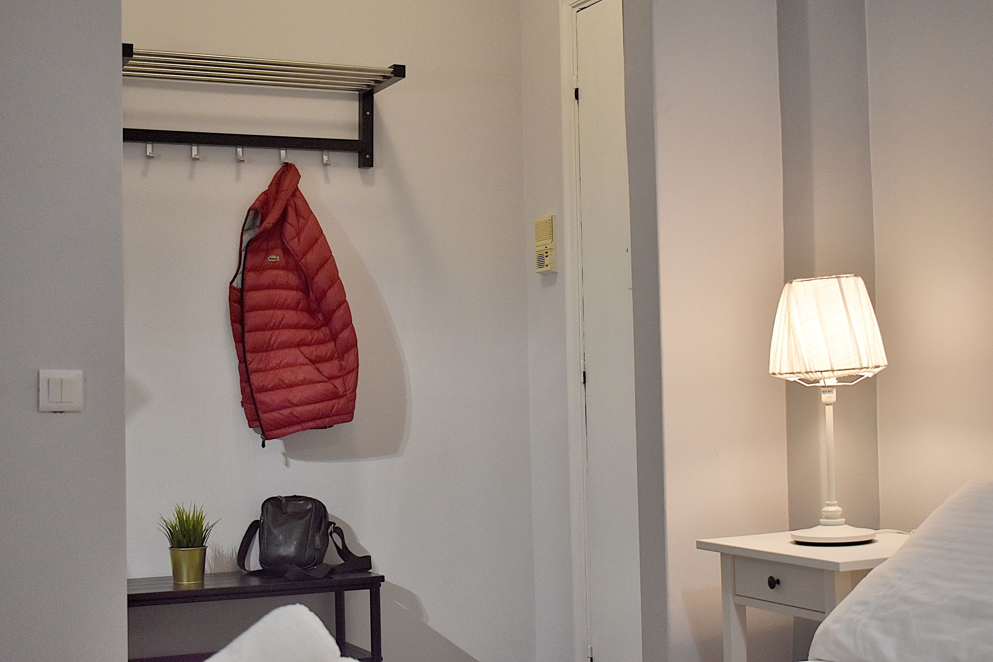 A hotel room with a bed, a bedside table with a lamp, a coat rack with a red jacket, a small plant, and a handbag.