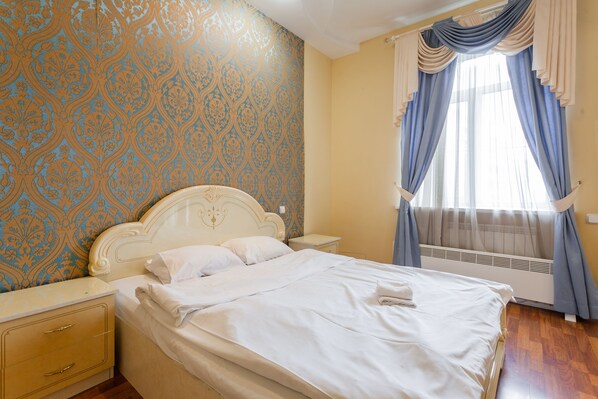 Cama bem arrumada com lençóis brancos, mesa de cabeceira de madeira e parede com papel de parede estampado em azul.