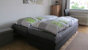 Un dormitorio con cama, mesita de noche, silla, radiador y ventana con vistas a casas.