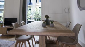 A dining area with a wooden table, six chairs, a hanging light fixture, and a view of a building through a window. - MAISON SALONIQUE 1930 (Thessaloniki)