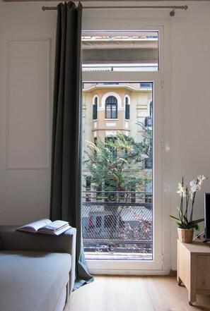 Una habitación con cama, ventana con vistas a edificios, una planta en maceta y una mesita.