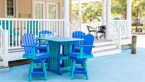 A covered outdoor seating area with a table and four chairs, a porch railing, and a view of a building with windows and doors.