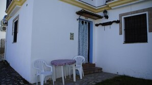 Un edificio blanco con una puerta azul, un pequeño patio con dos sillas de plástico blancas y un acceso empedrado de piedra.