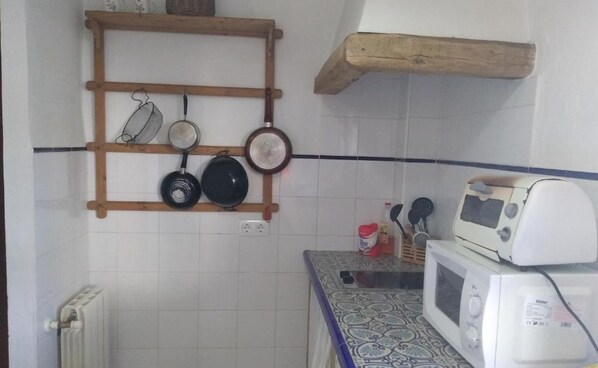 Una cocina con baldosas blancas, un estante de madera con ollas, un microondas y un mantel estampado.
