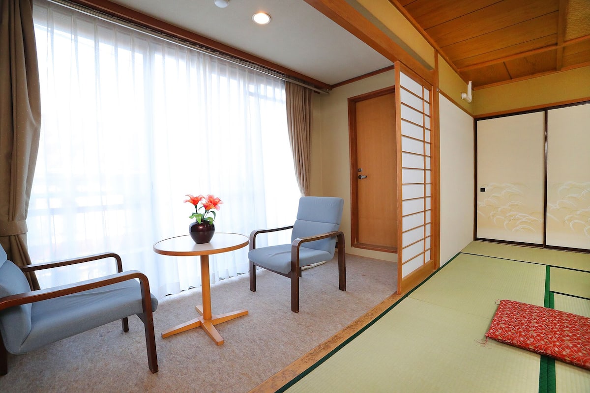 A traditional Japanese room with tatami flooring, a low table with a potted plant, two chairs, and sliding doors.