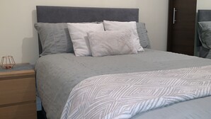 Un llit ben arropat amb un llençol estampat, una taula de nit de fusta amb una làmpada i un armari de color fosc.