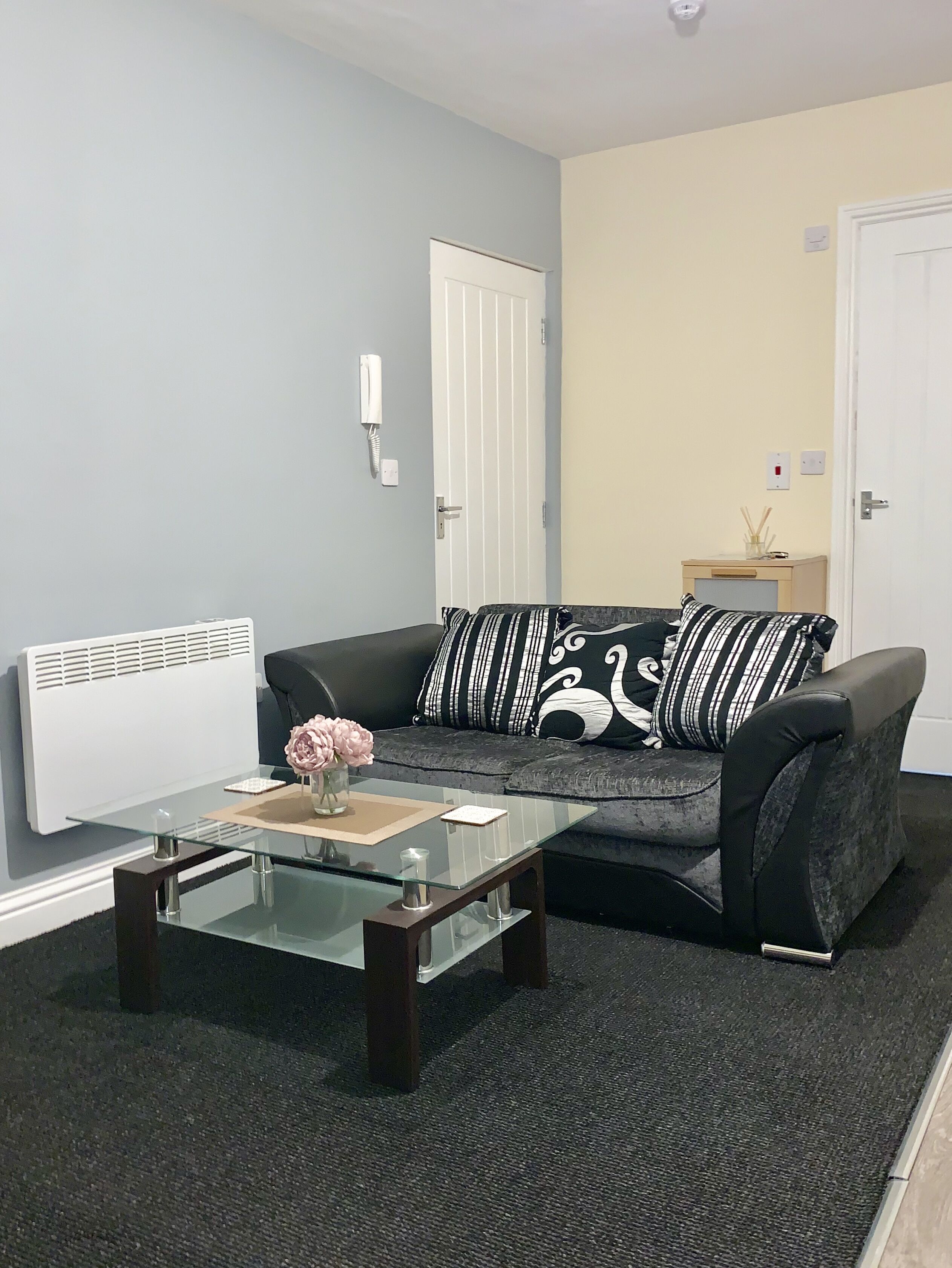 A living room with a grey sofa, black and white striped pillows, a glass coffee table with a vase of flowers, a white radiator, and a white door.
