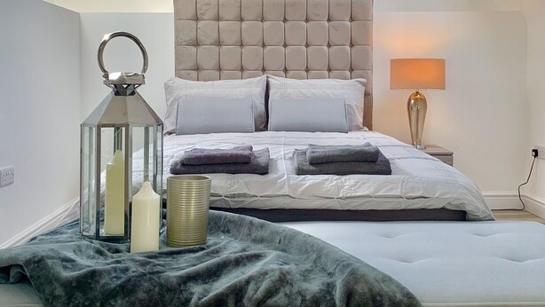 A bedroom with a high ceiling, a tufted headboard, a bed with white linens, a bedside lamp, and a skylight.