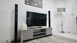 A modern living room with a flat-screen TV on a grey entertainment unit, a floor lamp, and a cityscape painting on the wall.