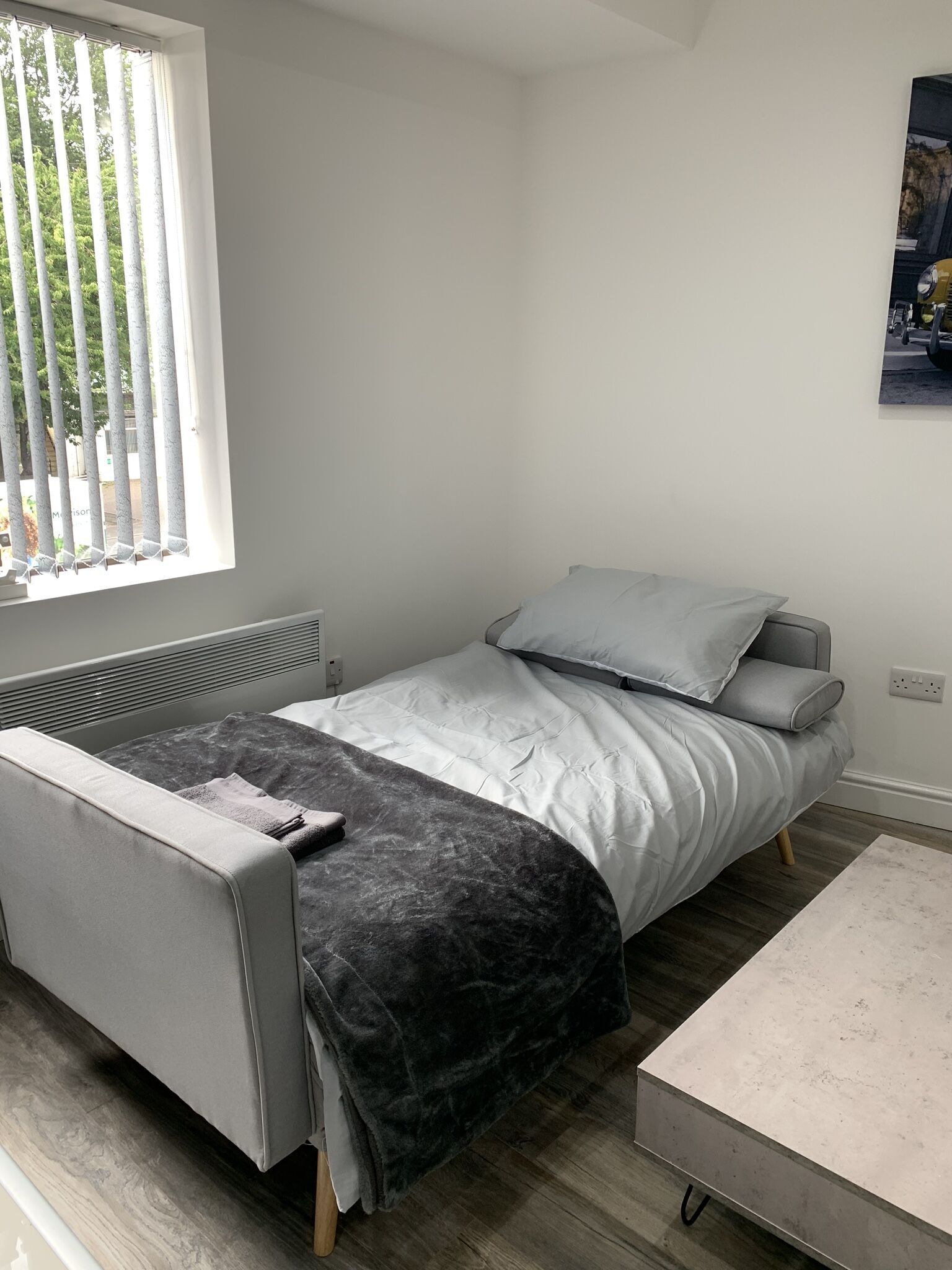 A single bed with a grey headboard and footboard, a grey blanket, and a grey pillow. There is a window with vertical blinds, a radiator, and a wall-mounted power outlet.