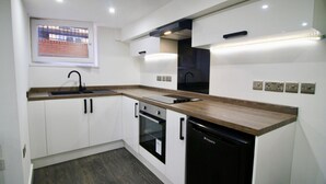 A modern kitchen with white cabinets, a wooden countertop, a black dishwasher, and a window with a view of a brick building. - StayHere 6 (Leeds)