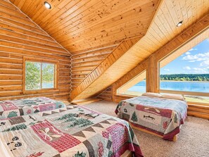 A log cabin bedroom with two beds, a sloped ceiling, and a window overlooking a lake. - Waters Edge* 9Br+loft, 5.5Ba, Boat Dock, Wifi (Island Park)