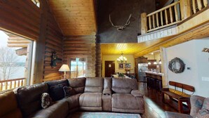 A log cabin interior with a large sectional sofa, wooden flooring, a chandelier, and a deer skull on the wall. - Waters Edge* 9Br+loft, 5.5Ba, Boat Dock, Wifi (Island Park)