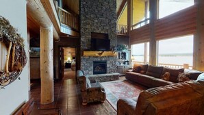 A spacious living area with a stone fireplace, leather sofas, and a large window overlooking a body of water. - Waters Edge* 9Br+loft, 5.5Ba, Boat Dock, Wifi (Island Park)