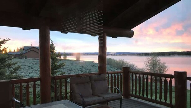 A covered deck with a wooden railing, a table, and a cushioned bench. - Waters Edge* 9Br+loft, 5.5Ba, Boat Dock, Wifi (Island Park)