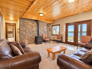 A cozy living room with leather sofas, a stone wall, a wood-burning stove, and a view of a lake through large glass doors. - Waters Edge* 9Br+loft, 5.5Ba, Boat Dock, Wifi (Island Park)