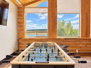 A foosball table in a room with large windows overlooking a lake and trees. - Waters Edge* 9Br+loft, 5.5Ba, Boat Dock, Wifi (Island Park)