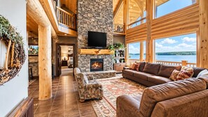 A spacious living room with a stone fireplace, leather sofas, and a large window overlooking a lake. - Waters Edge* 9Br+loft, 5.5Ba, Boat Dock, Wifi (Island Park)