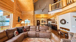 A log cabin interior with a large leather sectional sofa, a dining area, and a kitchen. - Waters Edge* 9Br+loft, 5.5Ba, Boat Dock, Wifi (Island Park)