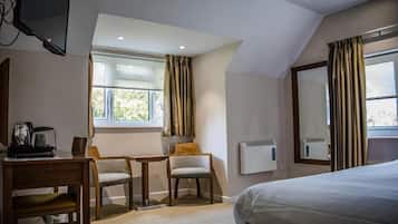 A hotel room with a bed, a desk, two chairs, a television, a window with curtains, and a wall-mounted heater.