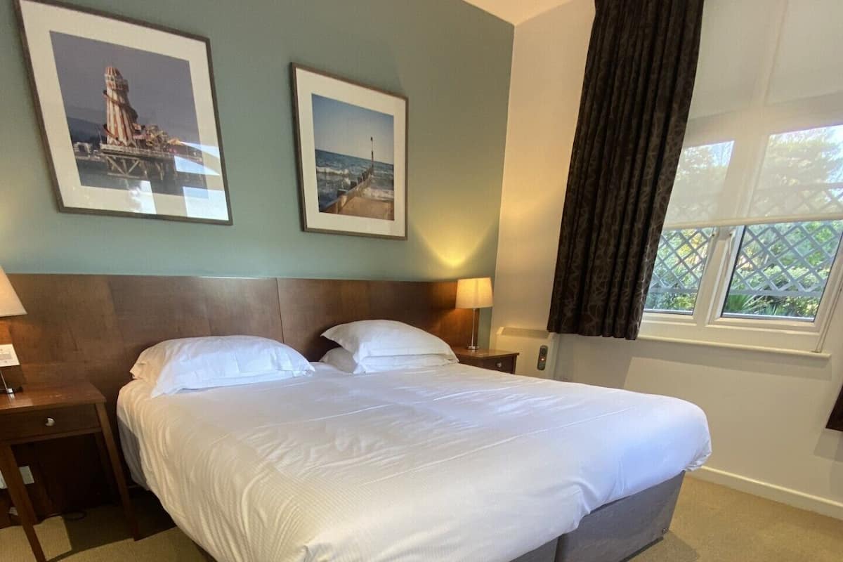 A neatly made bed in a hotel room with a headboard, two bedside tables, a lamp, and framed pictures on the wall.