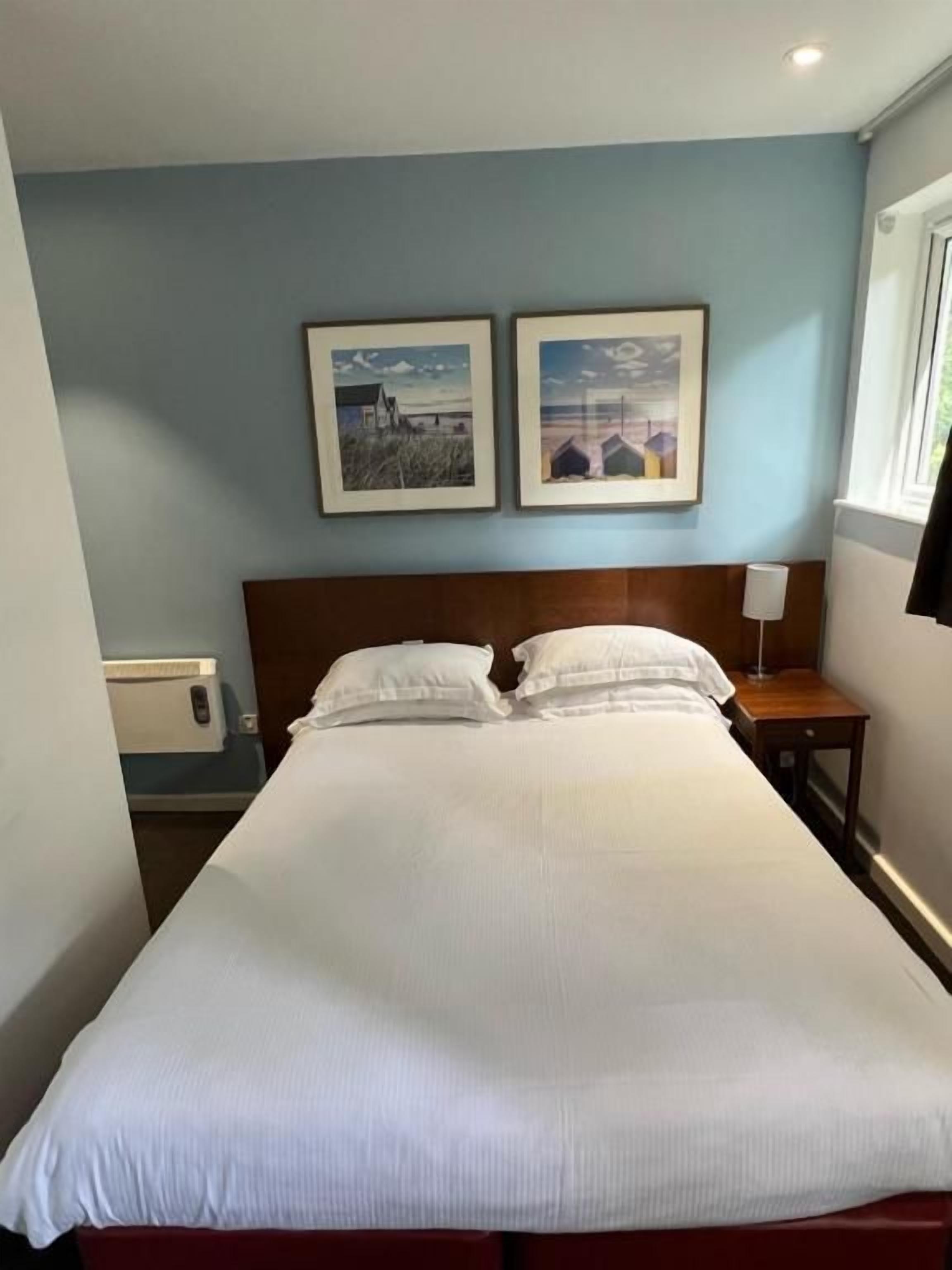 A hotel room with a large bed, two framed pictures on the wall, a bedside table, and a window.
