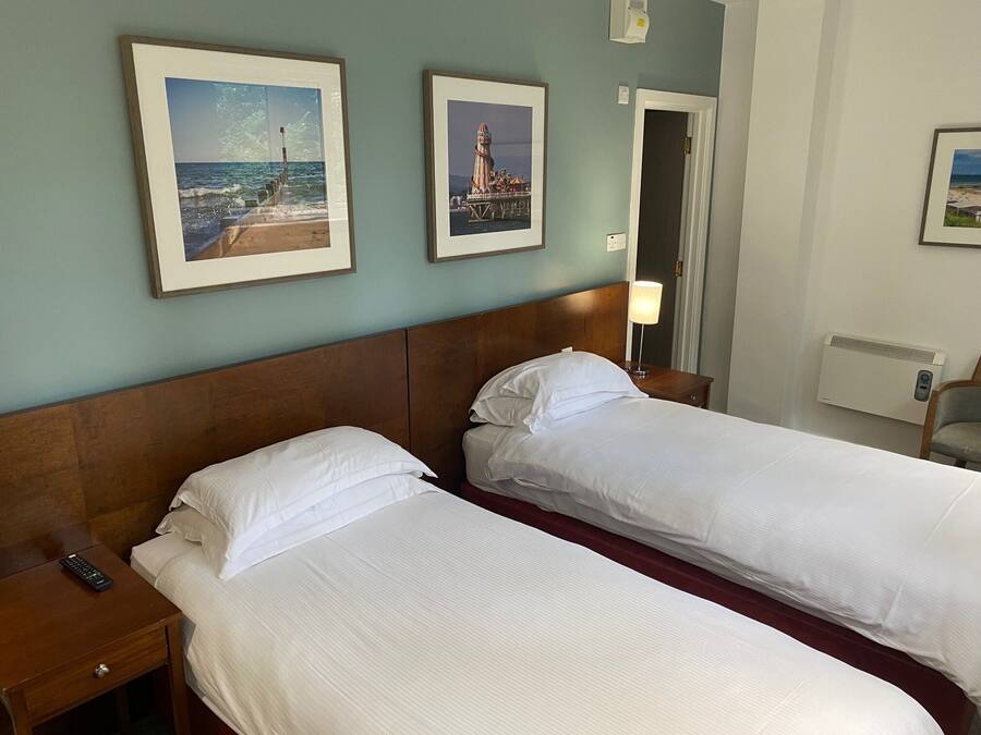Habitación de hotel con dos camas, cabecera de madera, mesita de noche, lámpara y cuadros en la pared.