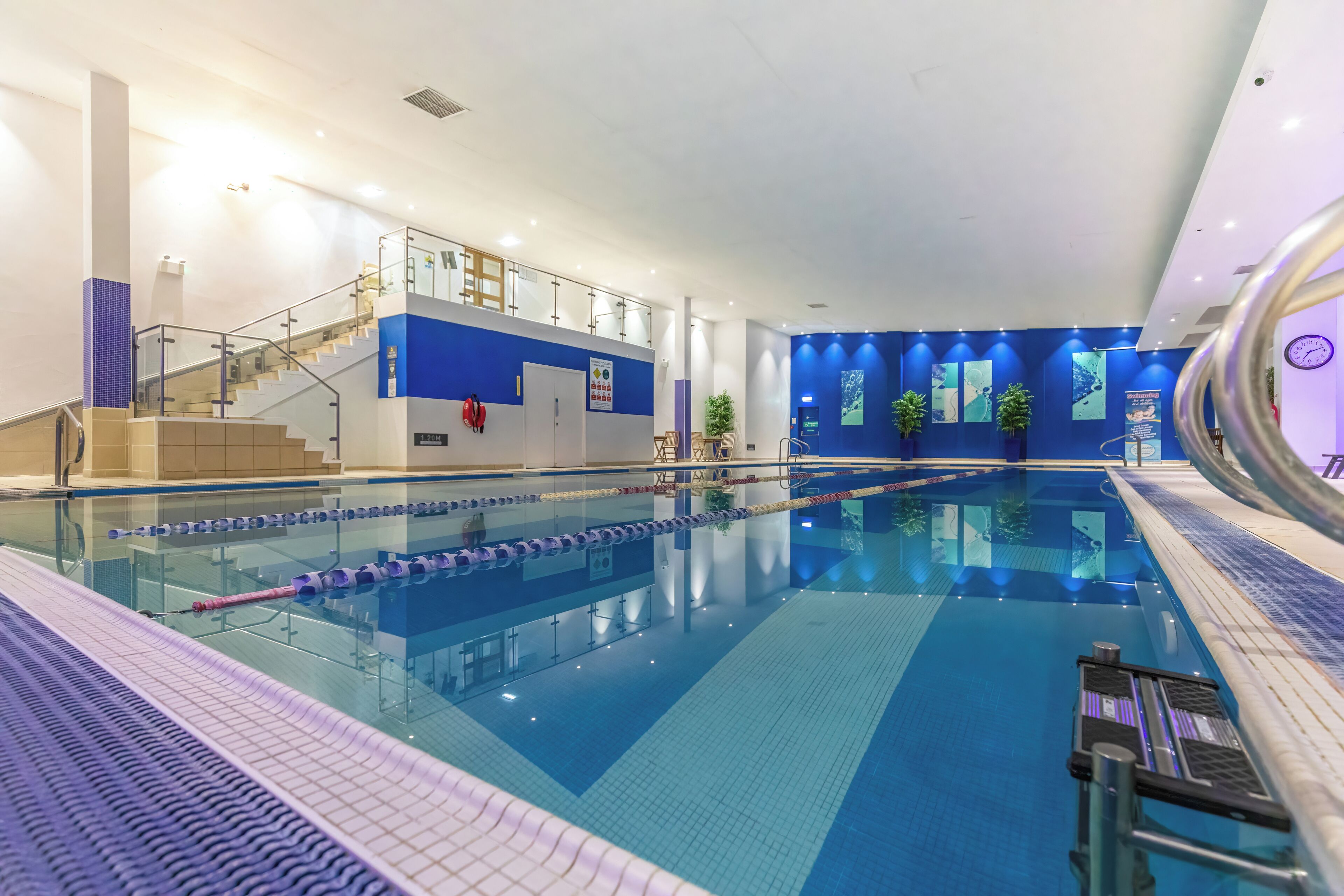 An indoor swimming pool with clear lanes, a staircase, and a blue wall with posters.