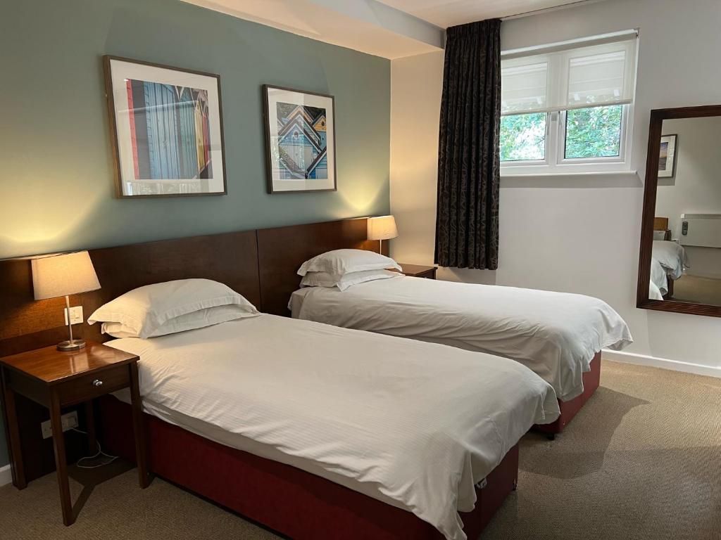 Habitación de hotel con dos camas, una mesita de noche, una lámpara, una ventana con cortinas y dos cuadros en la pared.