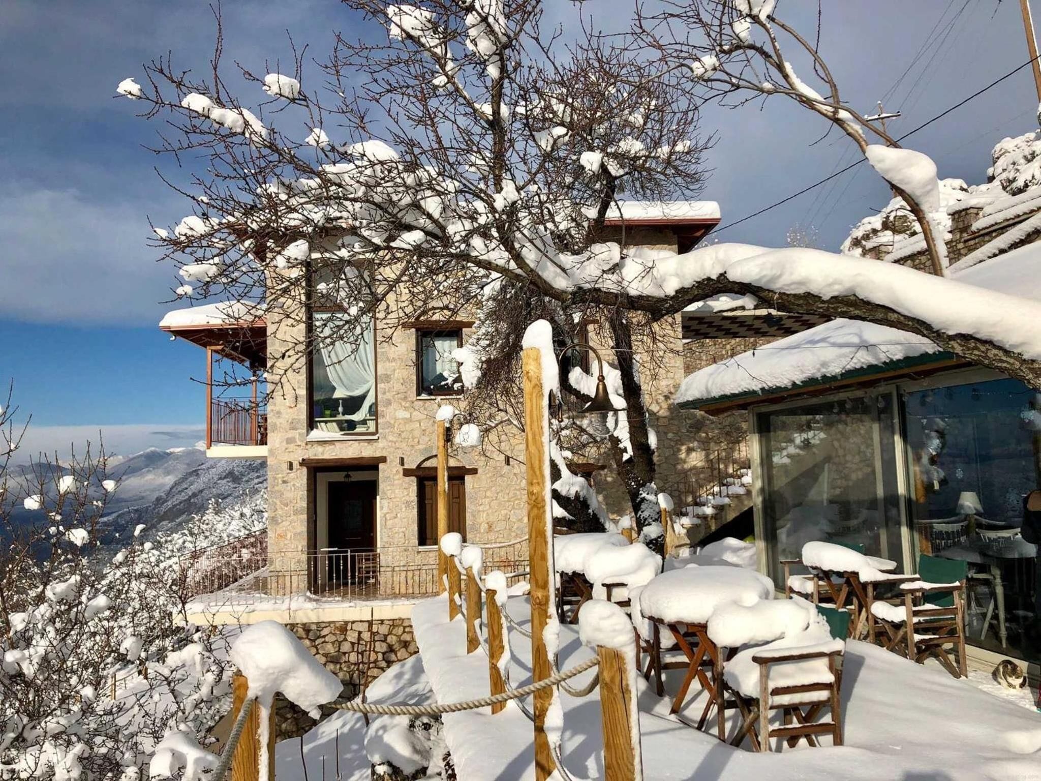 A stone building with a balcony, wooden chairs covered in snow, and a view of mountains.