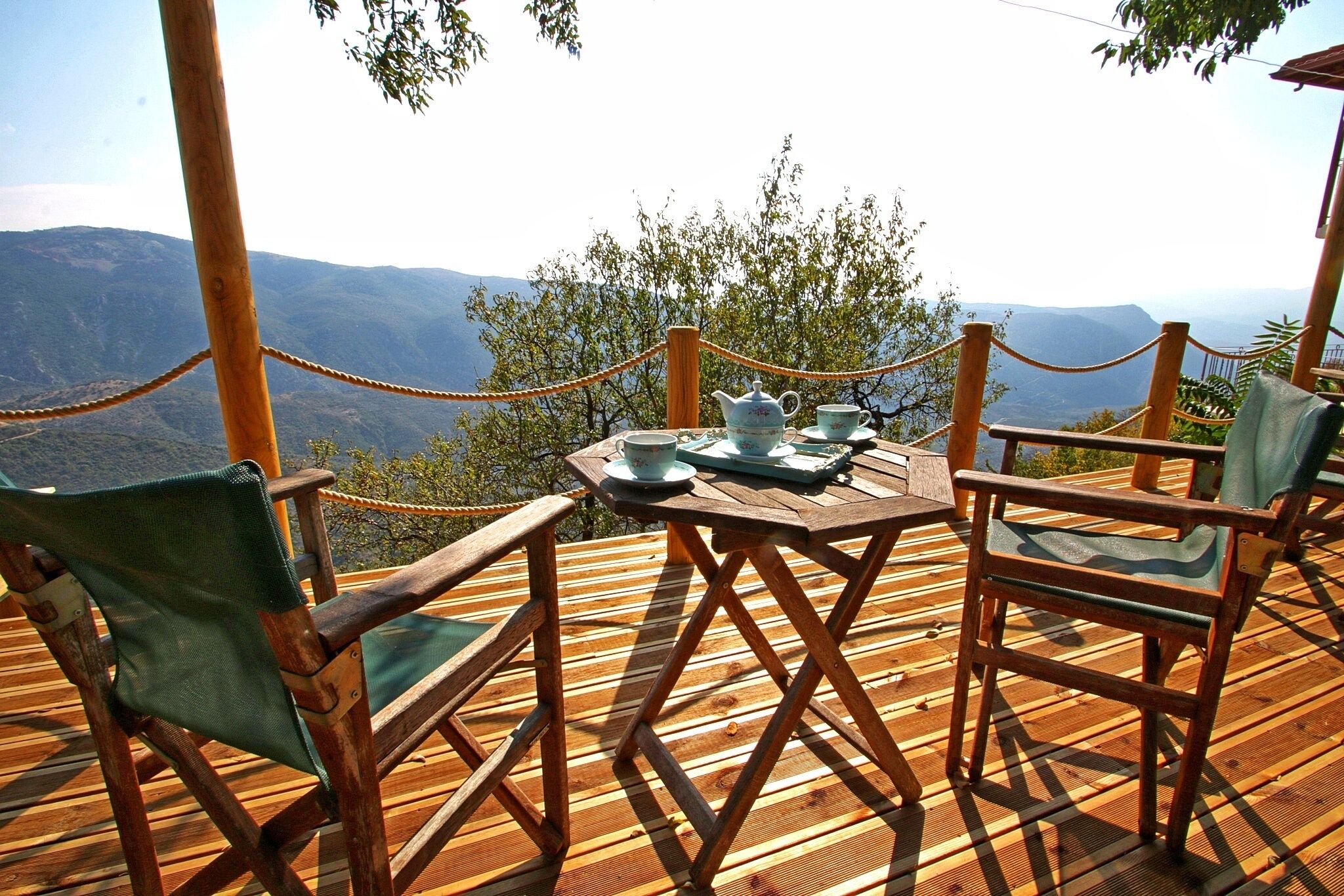 A wooden deck with a table set for tea, surrounded by chairs, overlooking a mountainous landscape.