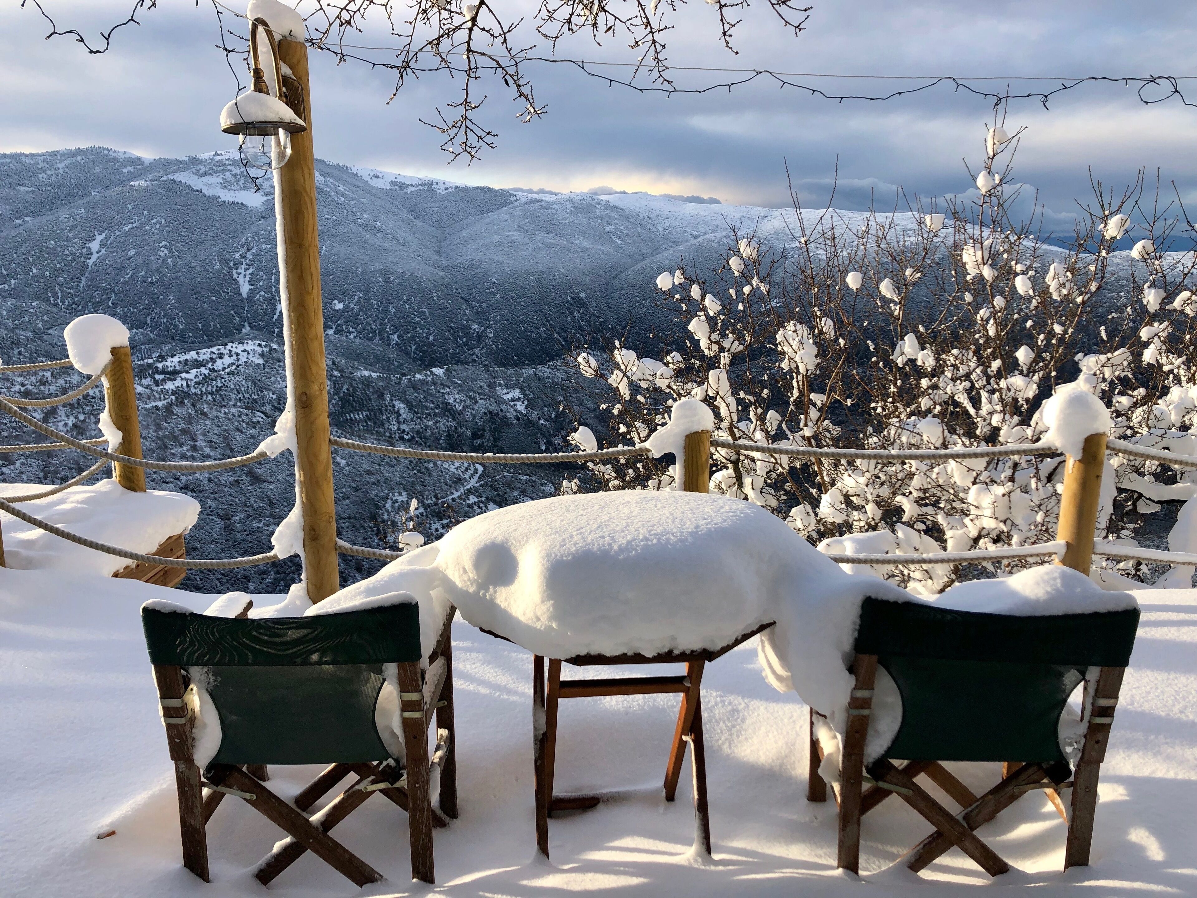 Ein mit Schnee bedeckter Außenbereich mit hölzernen Stühlen und einem Seilabfangnetz, mit Blick auf eine verschneite Berglandschaft.