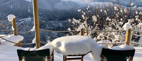 Ein mit Schnee bedeckter Außenbereich mit hölzernen Stühlen und einem Seilabfangnetz, mit Blick auf eine verschneite Berglandschaft.